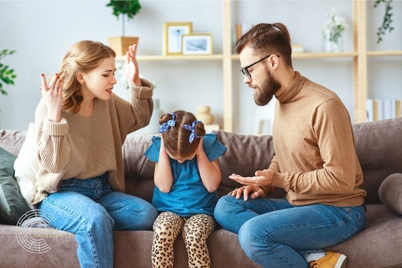 Divorcing parents quarrelling on sofa, with child in the middle with hands over ears